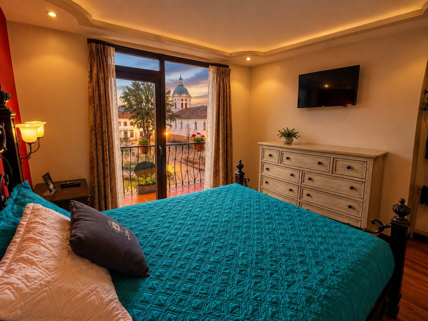 Habitación con cama de cubrecama turquesa, televisor y balcón con vista a una iglesia